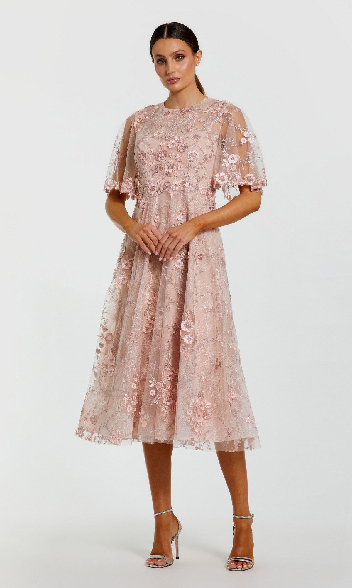 A woman stands in a blush Midi Wedding Guest Dress: Mac Duggal 69036 with floral-embroidered mesh and flutter sleeves, paired with silver heels against a plain background.